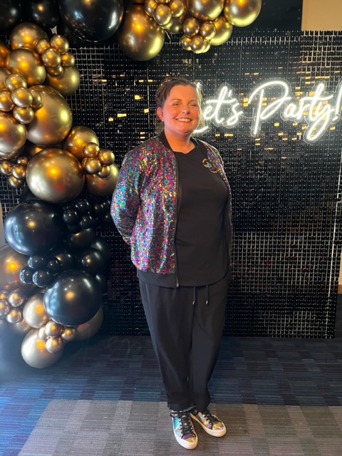 Lady in sequin jacket at a party with a sequin balloon wall behind her